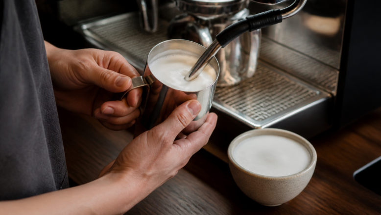 Barista schäumt Milch in einem Edelstahlkännchen auf [Bildinhalt mit KI erstellt]